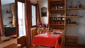 A dining area with a red tablecloth, wooden chairs, a wooden bench, and a bookshelf with various kitchen items. - Cozy and bright apartment, 2 rooms, near slopes, 4 people (Pralognan-la-Vanoise)
