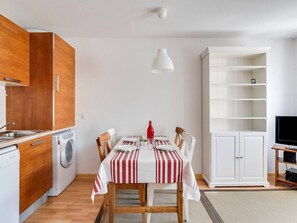 Uma cozinha compacta com armários de madeira, uma máquina de lavar, uma mesa de jantar com toalhas listradas e um armário branco com prateleiras.