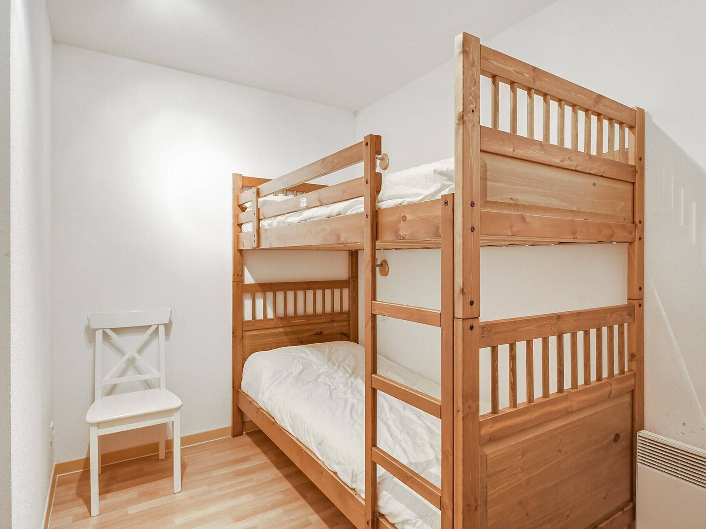 A bunk bed with a wooden frame and white bedding, a white chair, and a radiator on the floor.