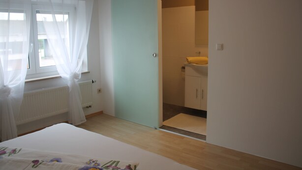 A bedroom with a bed, a window with sheer curtains, a radiator, and a bathroom with a sink and cabinet. - Appartement- Steinkirchner (Straubing)