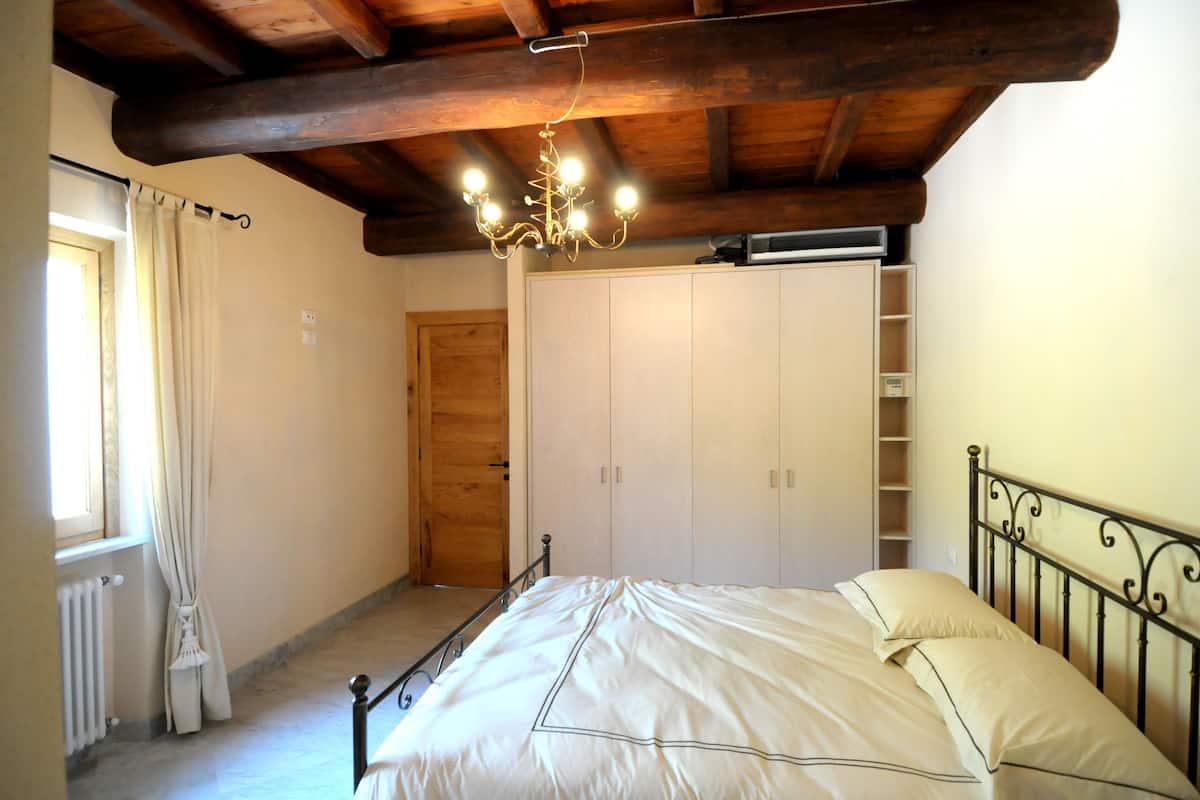 Une chambre avec un plafond en bois, un lit avec une literie blanche, un lustre, une fenêtre avec des rideaux et une armoire.