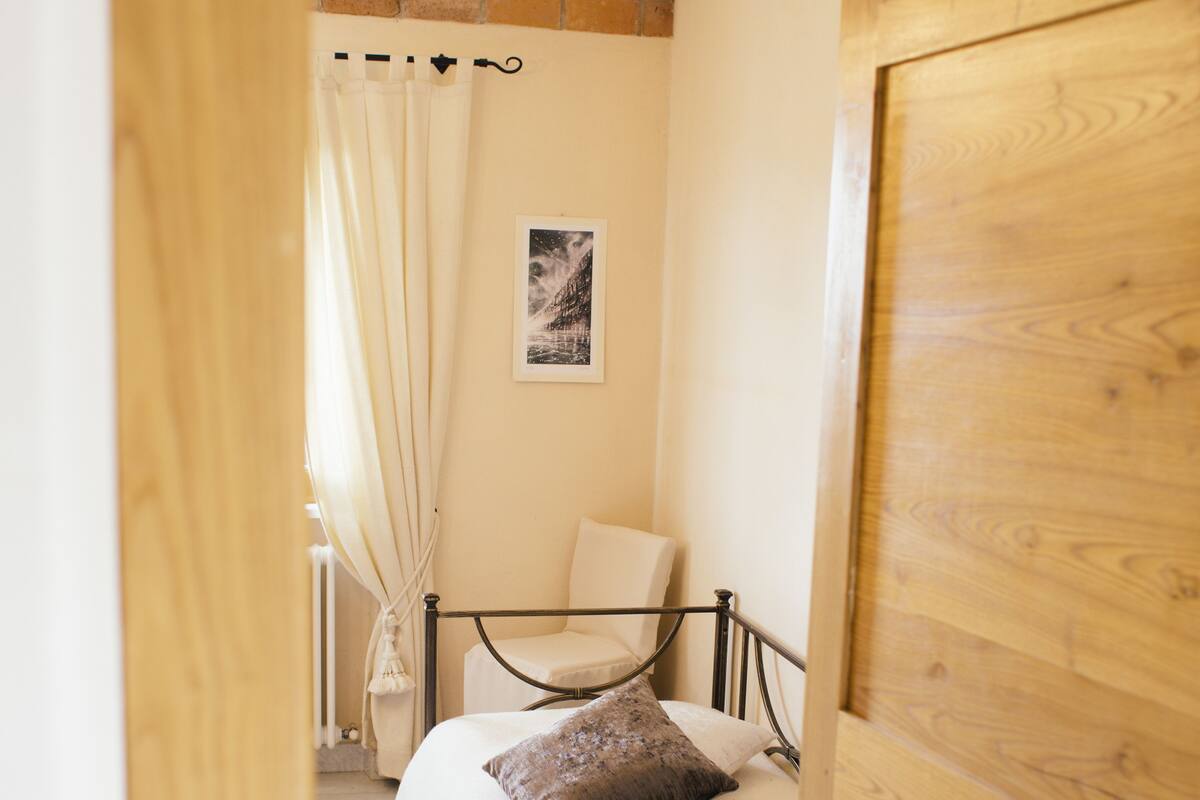 Une chambre avec une porte en bois, un lit avec une literie blanche, une chaise et une fenêtre avec des rideaux.