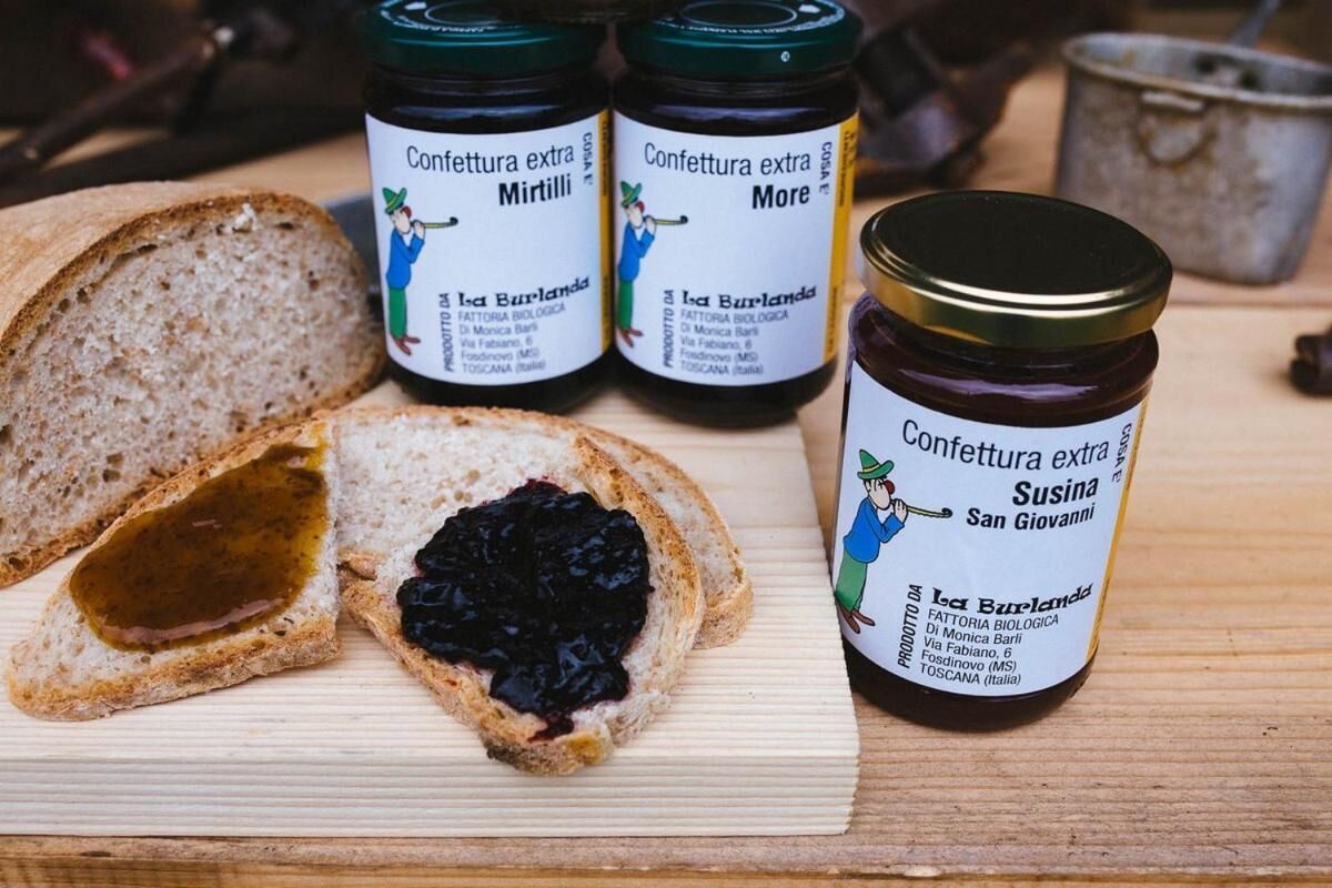 A wooden board with slices of bread, two jars of extra confettura, and a jar of Susina.