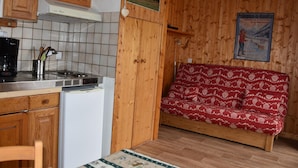 A compact kitchen with wooden cabinets, a refrigerator, and a stove. There is a wooden bench with a patterned cushion in the living area. - Cozy studio near shops, south balcony (Pralognan-la-Vanoise)