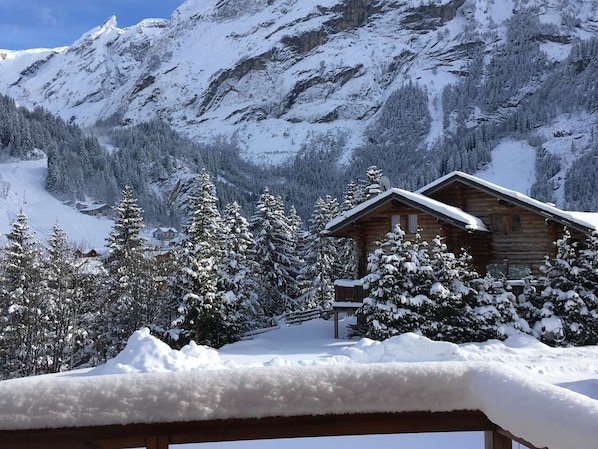 Une cabane en bois, dont le toit est recouvert de neige, entourée d’arbres chargés de neige et avec des montagnes en arrière-plan.