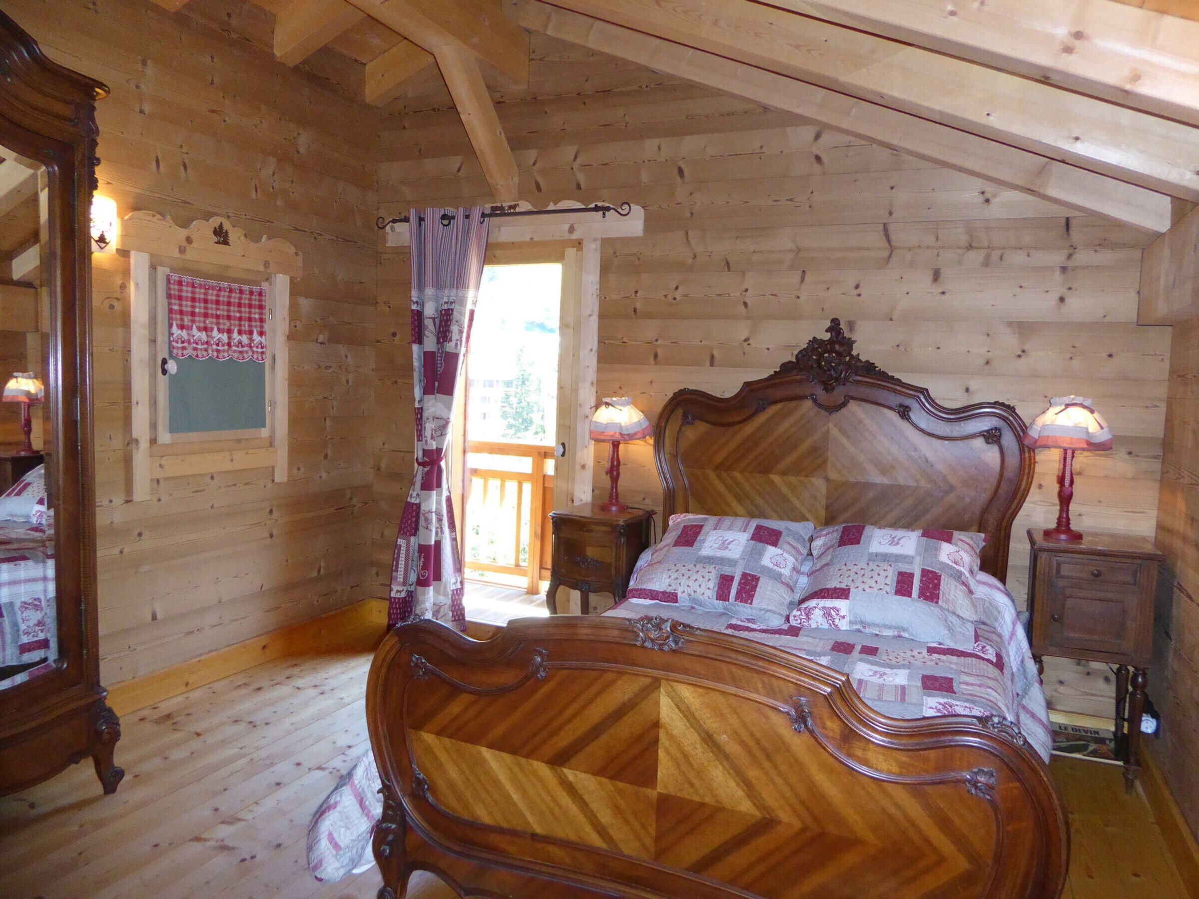 A wooden cabin bedroom with a large bed, two bedside tables with lamps, a window with curtains, and a mirror on the left wall.