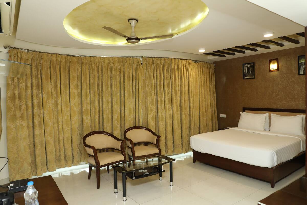 A hotel room with a bed, two chairs, a glass coffee table, a ceiling fan, and curtains.