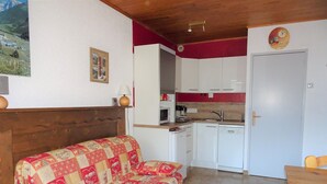 Ein gemütliches Zimmer mit einem hölzernen Kopfteil, einer gesteppten Couch, einem kleinen Tisch, einer Küche mit weißen Schränken und einer roten Akzentwand.