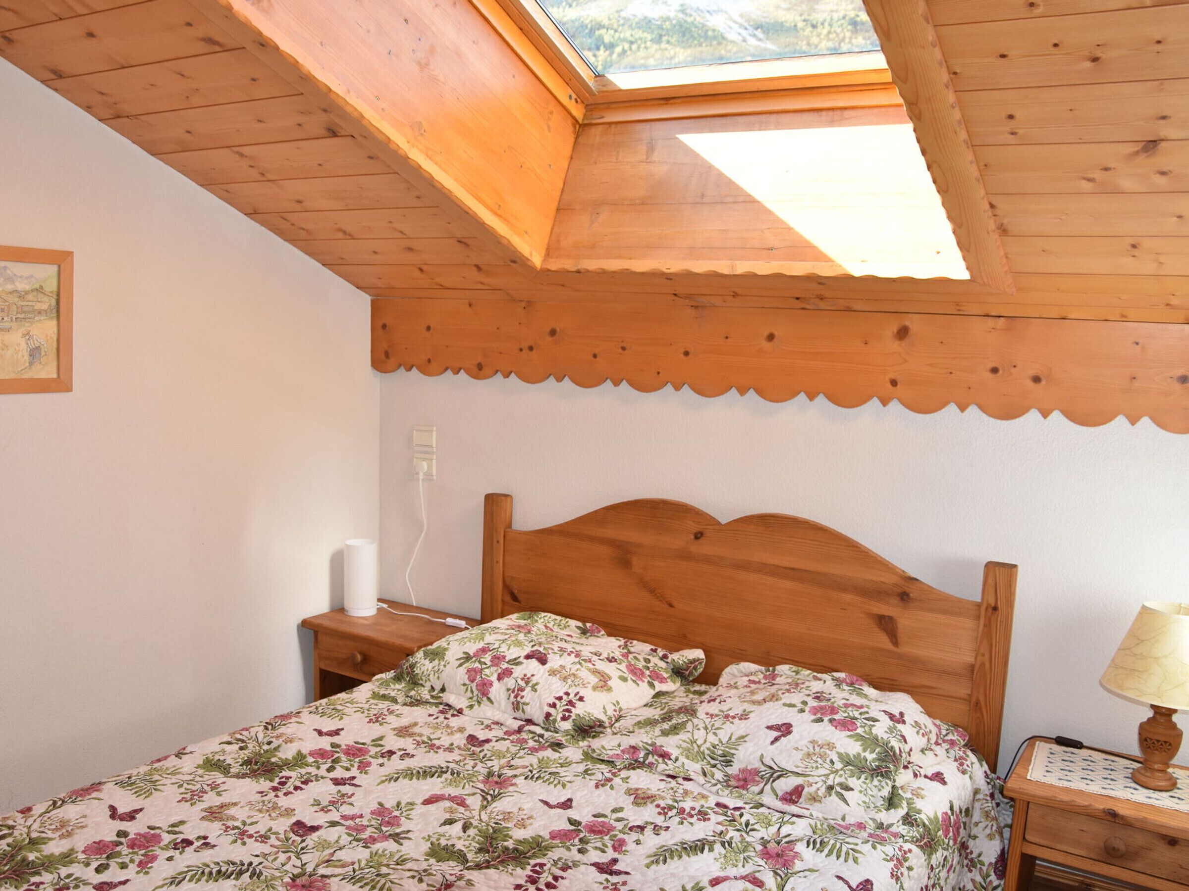 Quarto com teto de madeira, cama com roupa de cama floral, luminária de cabeceira e mesa de apoio.