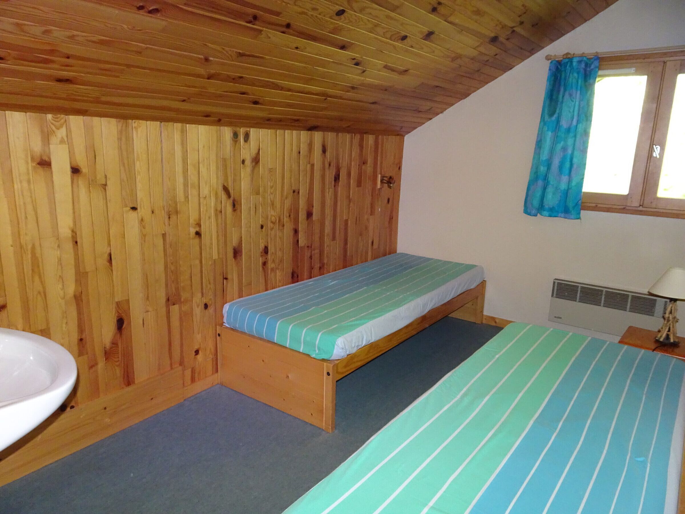 A small, single-occupancy room with a wooden ceiling and walls, a window with a blue curtain, a bed with striped bedding, a bedside table with a lamp, and a sink.