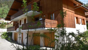 Een houten chalet met twee verdiepingen, met een balkon op de bovenverdieping, omringd door groen.