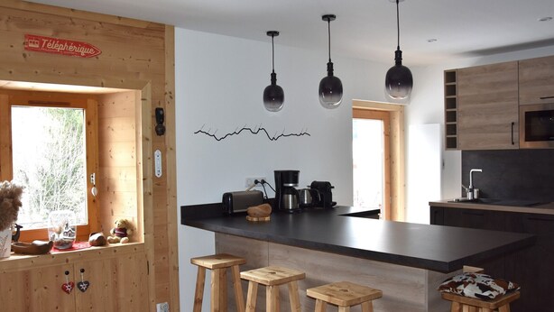 A modern kitchen with wooden accents, a central island with stools, pendant lights, and a visible refrigerator.