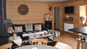 A cozy living room with a fireplace, wooden walls, a sofa with patterned cushions, a dining table, and a television mounted on the wall.
