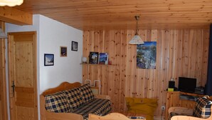A cozy room with wooden walls and furniture, including a sofa, a yellow chair, and a small table. There is a television on a wooden stand and a painting on the wall.