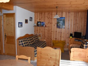 A cozy room with wooden walls and furniture, including a sofa, a yellow chair, and a small table. There is a television on a wooden stand and a painting on the wall.