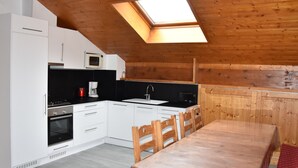 Une cuisine compacte avec des meubles blancs, un four à micro-ondes, une cuisinière et un évier. Il y a une lucarne au-dessus de l’évier et une table à manger avec des chaises.