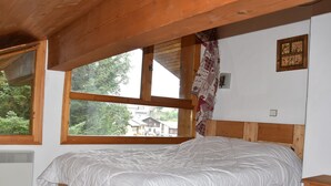 Une chambre avec une grande fenêtre, des poutres en bois au plafond, un lit avec une literie blanche, une table de chevet en bois et un interrupteur fixé au mur.