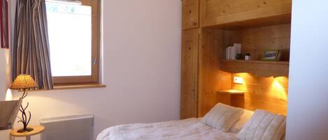 Une chambre à coucher avec un lit, une fenêtre avec des rideaux, un radiateur, une lampe et une armoire en bois.