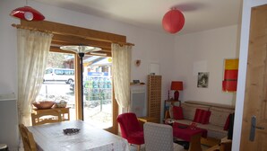 A dining area with a table and chairs, a sofa, a red lamp, and a view of the outside. - Pleasant 3-room apartment with garden, close to the center (Pralognan-la-Vanoise)