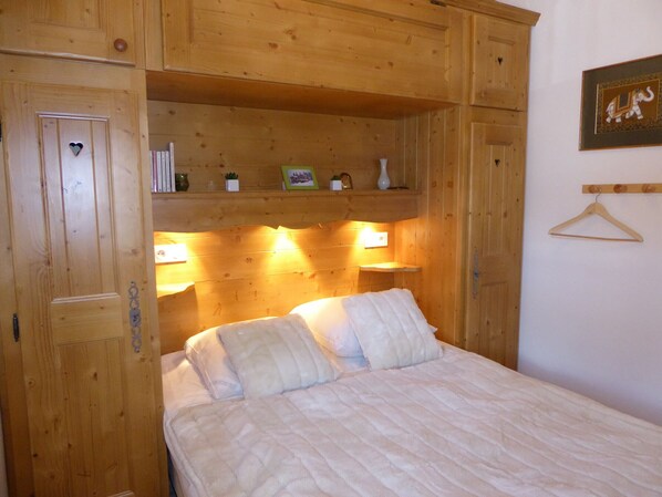 A wooden room with a bed, shelves with books and decorative items, and a wall-mounted light fixture.
