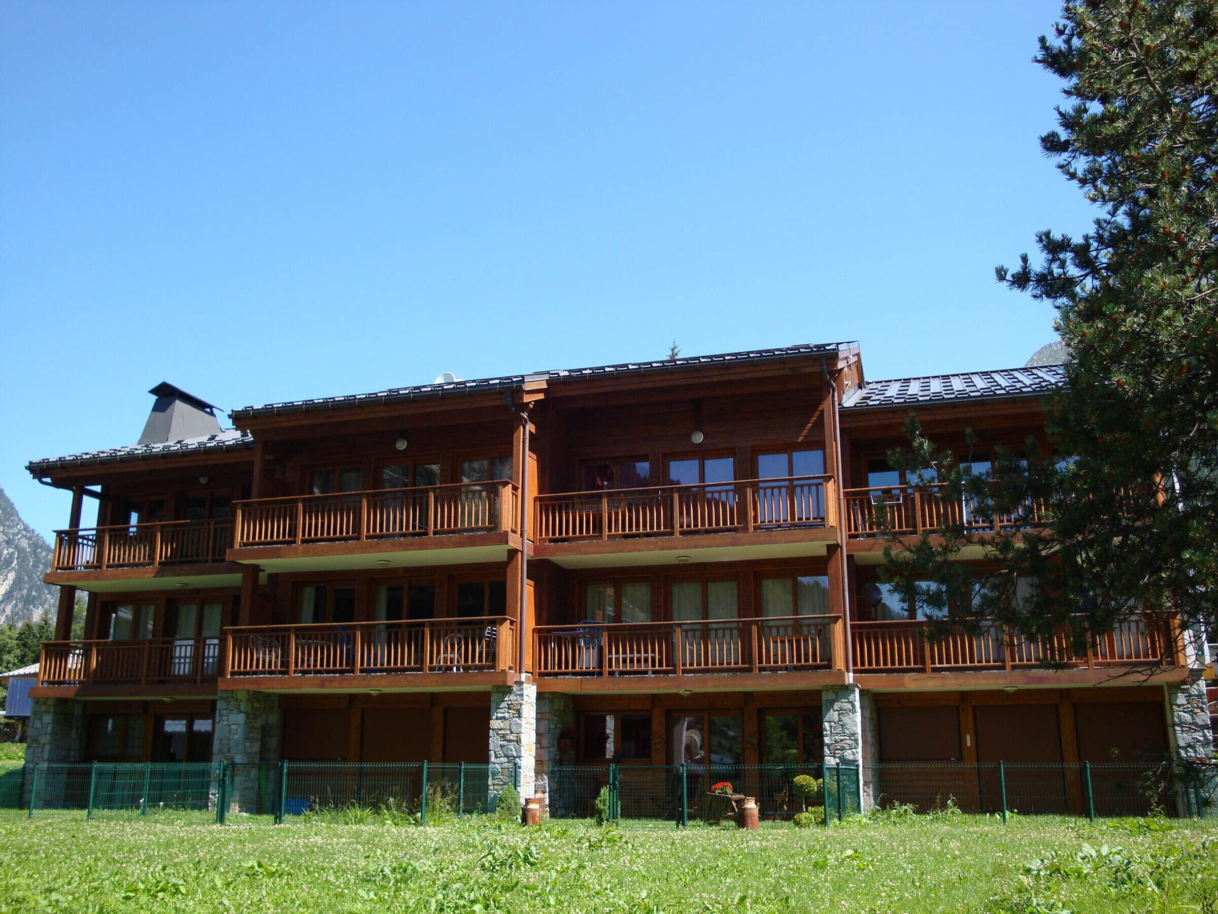 A multi-story wooden building with balconies, stone pillars, and a grassy foreground.