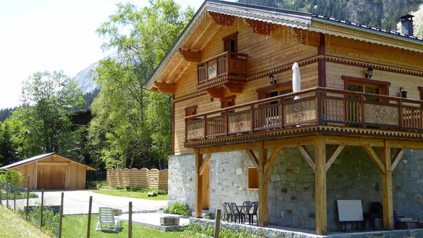 Een houten chalet met balkon, stenen fundering en uitzicht op het omliggende bos en de bergen.