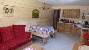 Een gezellige kamer met houten details, een bank, een tafel met tafelkleed, een kroonluchter, een keukenhoek met houten kastjes en een magnetron.