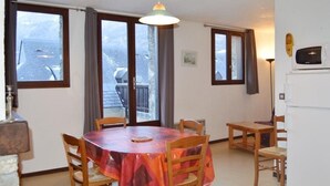 A dining area with a round table covered by a red and orange tablecloth, surrounded by wooden chairs. There are two windows with curtains, a ceiling light, and a white refrigerator in the background. - Duplex T4 for 7 with terrace and parking in Esquièze-Sère (Esquièze-Sère)