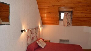 A bedroom with a wooden ceiling, a bed with a red blanket, a small wooden stool, and a window with floral curtains. - Duplex T4 for 7 with terrace and parking in Esquièze-Sère (Esquièze-Sère)