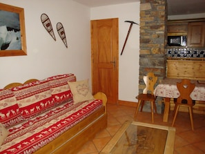 A cozy living room with a wooden sofa, a small table, and chairs. A wooden door and a stone wall are visible in the background.