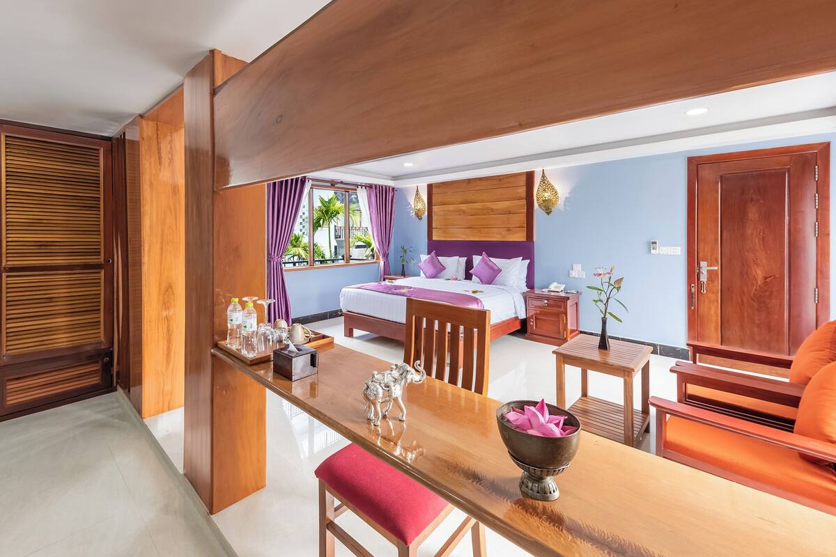 Ein Hotelzimmer mit Bett, Schreibtisch aus Holz, Stuhl, Nachttisch und Blick ins Freie.