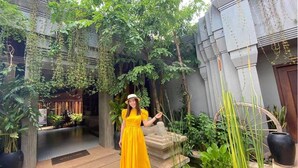 A woman in a yellow dress standing in a courtyard with a fountain, surrounded by greenery and a stone pathway. - Cheata Residence (Siem Reap)