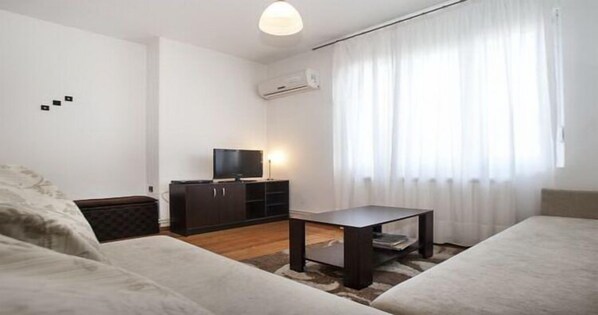 A modern living room with a sofa, a coffee table, a TV on a cabinet, and a large window with curtains. - Apartment Rubin (Belgrade)