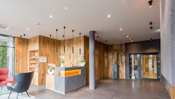 Un área de recepción moderna con un mostrador de madera, sillas negras y una entrada por puerta de cristal.