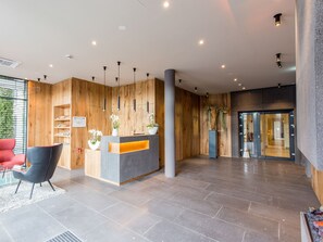 A modern reception area with a wooden reception desk, black chairs, and a glass door entrance. - monte mare HOTEL Andernach (Andernach)