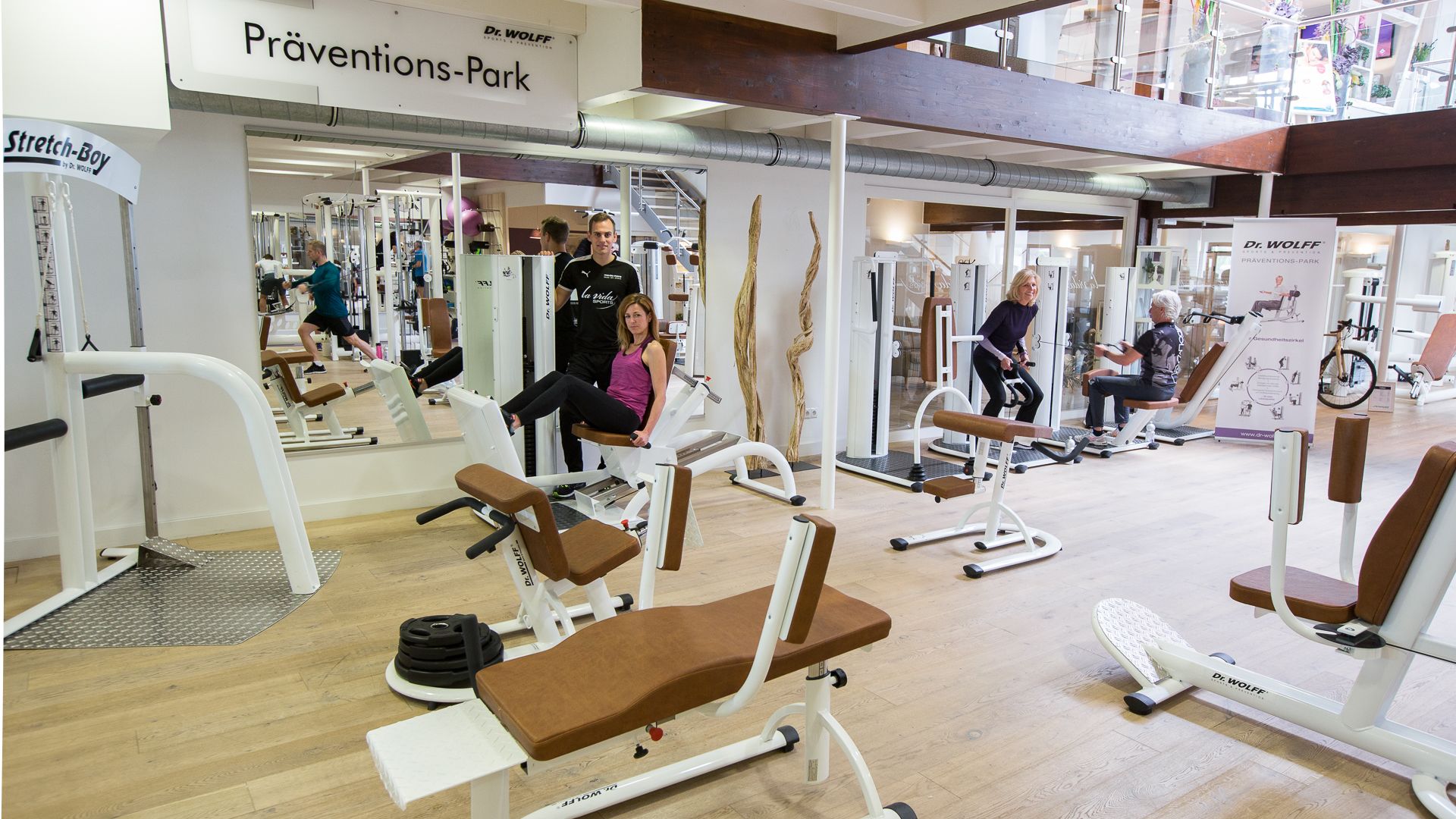 Una palestra con diverse attrezzature per esercizi, tra cui panche e macchinari per pesi, e un cartello per Dr. Wolff's Prevention Park.