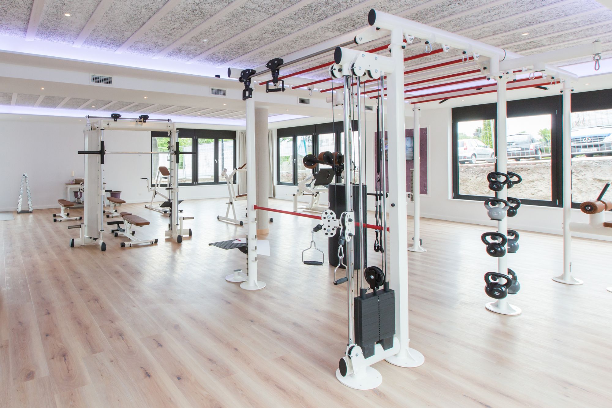 A spacious gym with various exercise equipment, large windows, and a wooden floor.