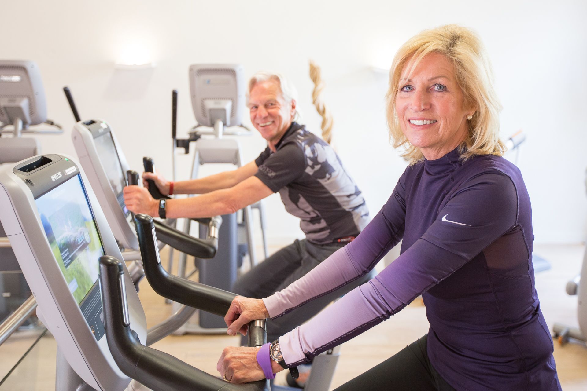 Two people using stationary bikes in a gym with exercise equipment in the background.