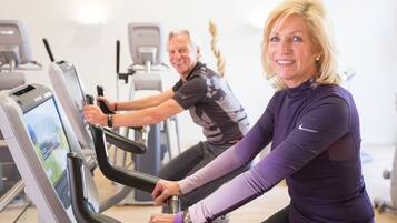 Two people using stationary bikes in a gym with exercise equipment in the background.