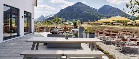 Une terrasse extérieure avec une table et des bancs en bois, entourée de chaises et de parasols jaunes, offrant une vue sur un paysage montagneux.