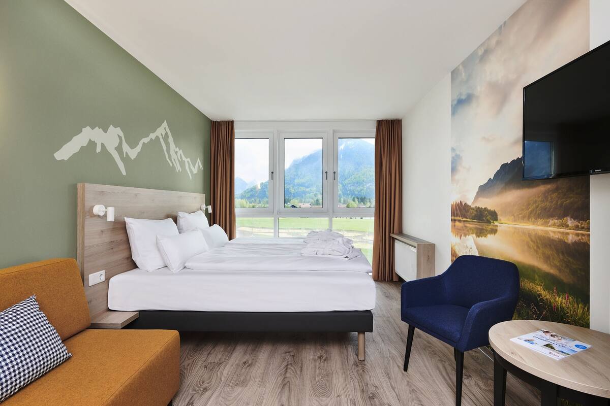 A hotel room with a bed, a sofa, a chair, a table, a TV, and a large window with a mountain view.