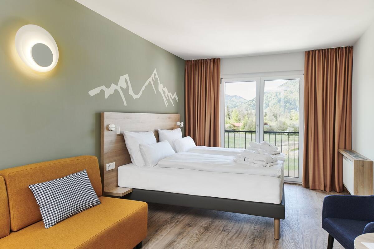 A modern hotel room with a bed, a sofa, a chair, a wall-mounted light, and a view of the outdoors.