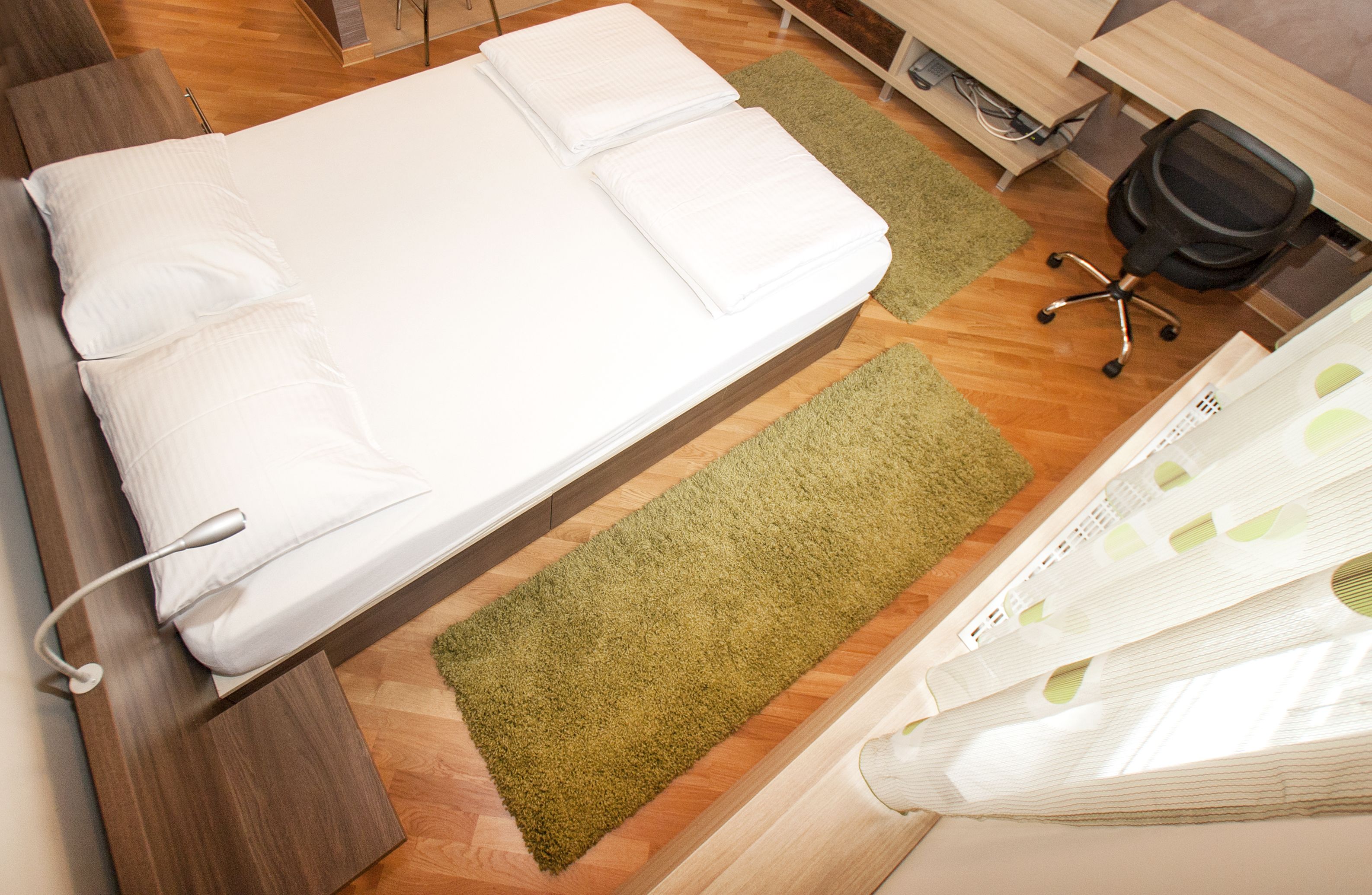 Un lit bien fait, avec des oreillers et un tapis vert, un bureau en bois avec une chaise, et une fenêtre avec des rideaux.
