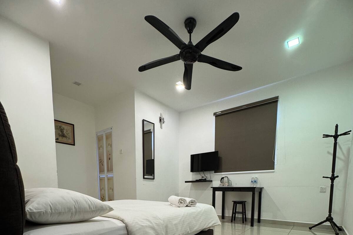 A modern hotel room with a bed, a desk with a computer, a standing lamp, and a ceiling fan.