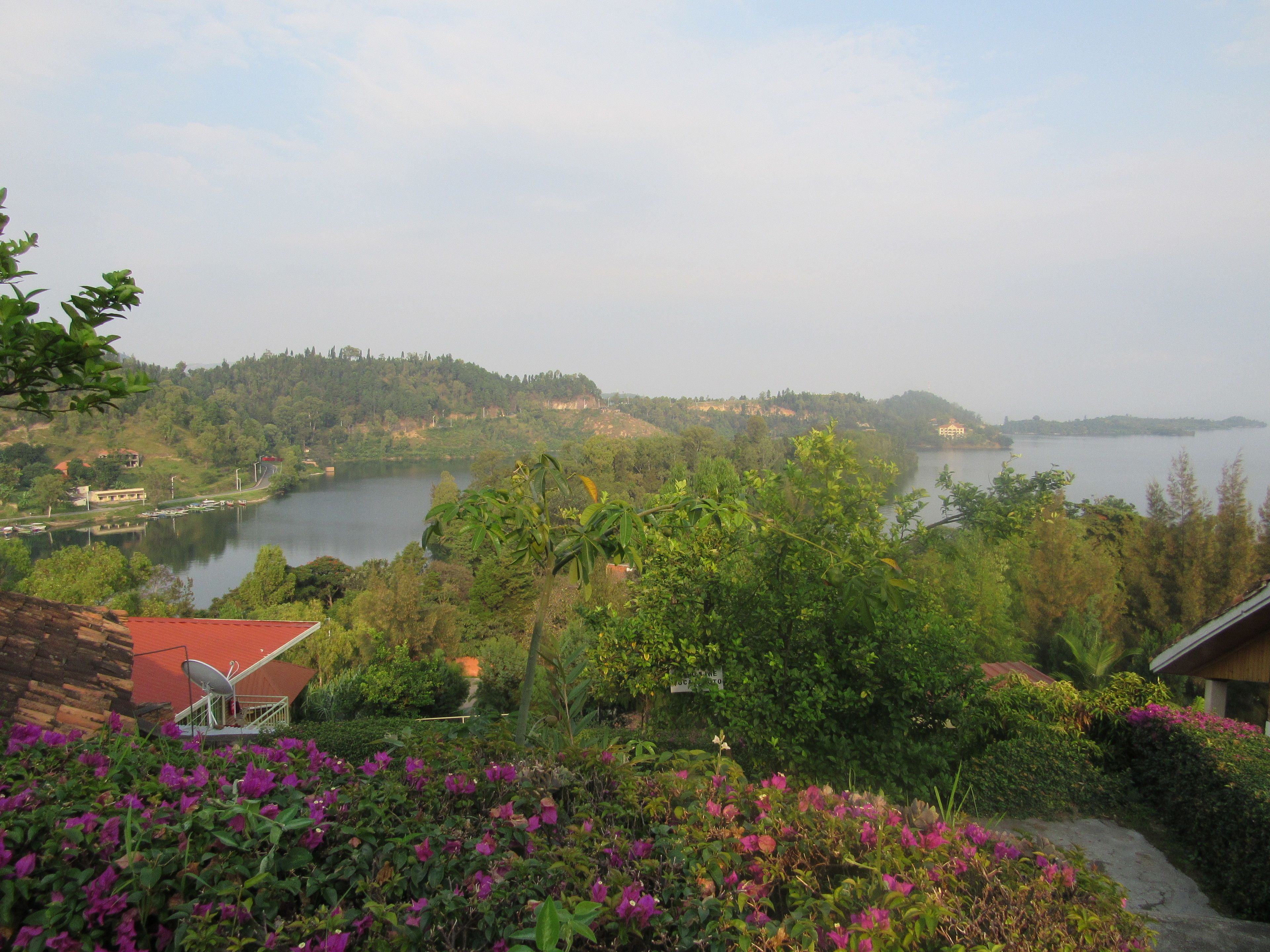 A view of a lake surrounded by greenery and hills. There is a building with a red roof in the foreground.