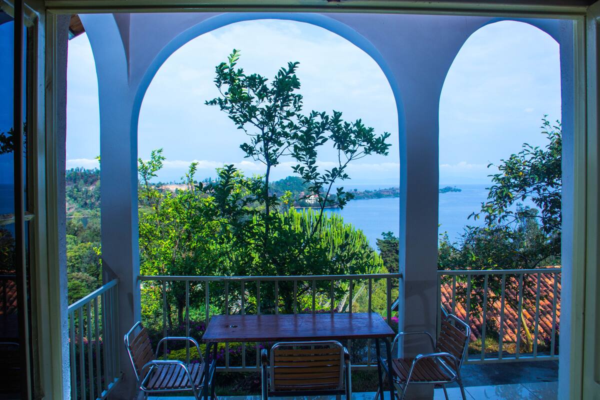 Un balcon avec une table et des chaises, offrant une vue panoramique sur un paysage verdoyant composé d’arbres et d’une étendue d’eau.