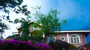 A traditional brick house with a tiled roof, surrounded by lush greenery and vibrant purple flowers. - Rebero Kivu Hotel (Bwishyura)