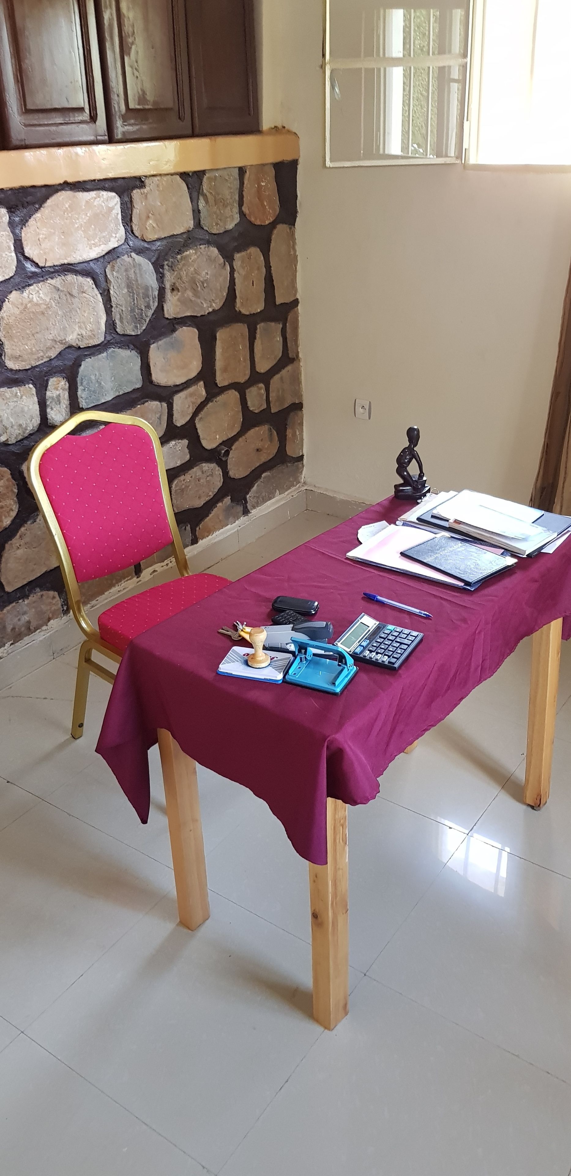 A room with a stone wall, a wooden door, a window, a table with a red tablecloth, a chair, and various items on the table.