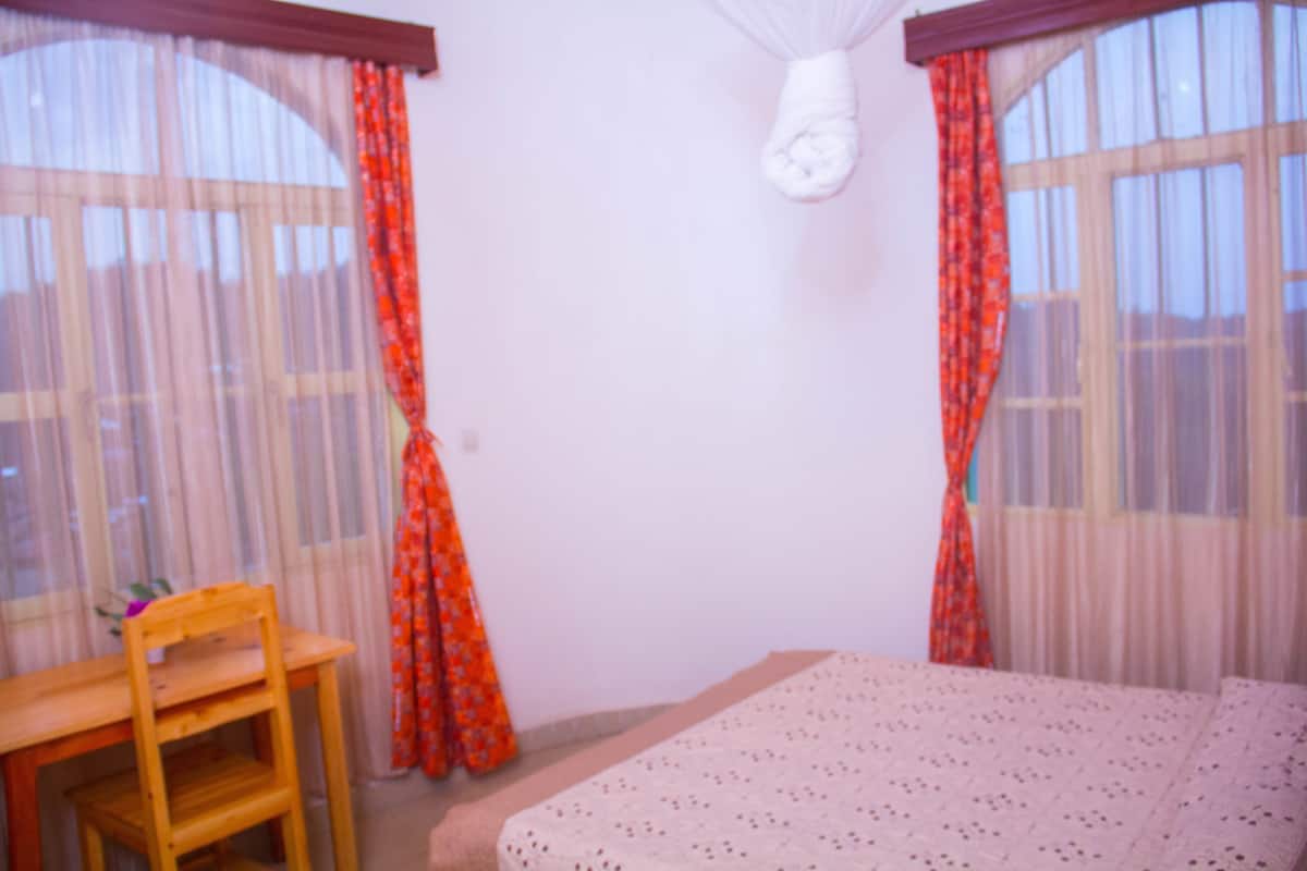 Une chambre à coucher avec un lit, une chaise en bois et une fenêtre ornée de rideaux légers.