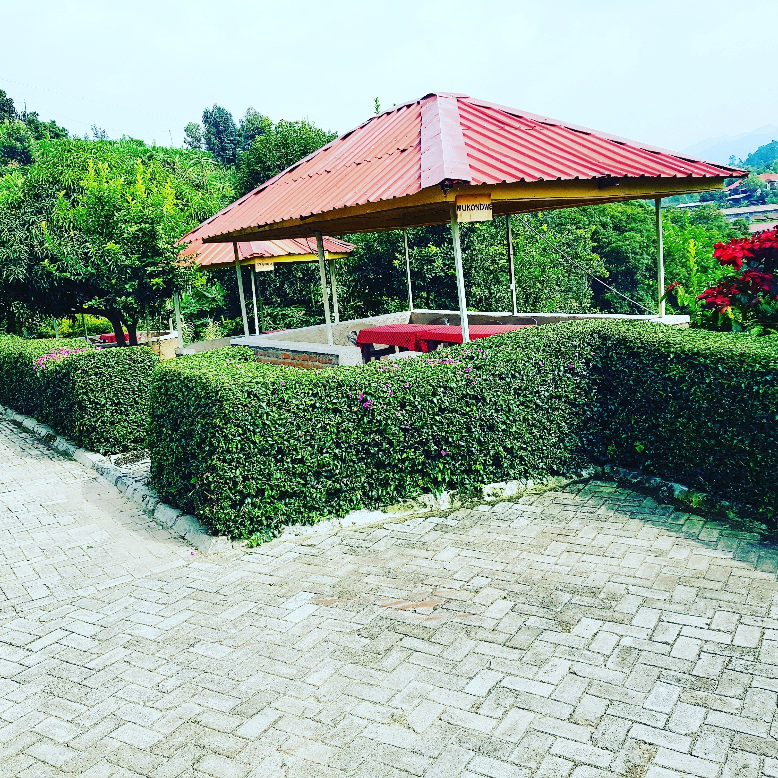 A covered pavilion with a red roof, surrounded by well-maintained hedges and a paved walkway.